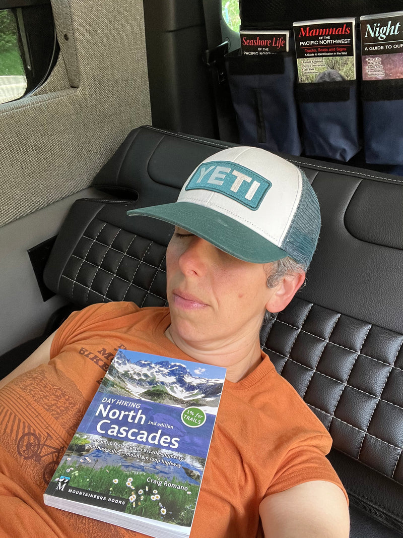 An adventurer naps in an RV rental, with a "Day Hiking North Cascades" book on their chest & nature guides nearby. — photo 15