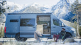 Experience winter van life with this RV rental. A bearded man sips coffee outside his blue camper van, surrounded by snowy mountains.