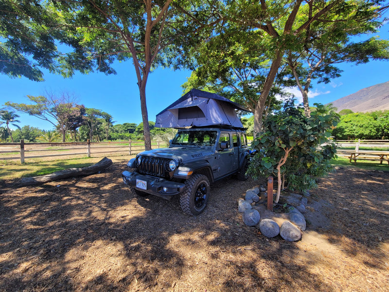 A grey Jeep Gladiator RV rental with a rooftop tent, parked on a dirt lot under large shade trees, ready for adventure. — photo 1