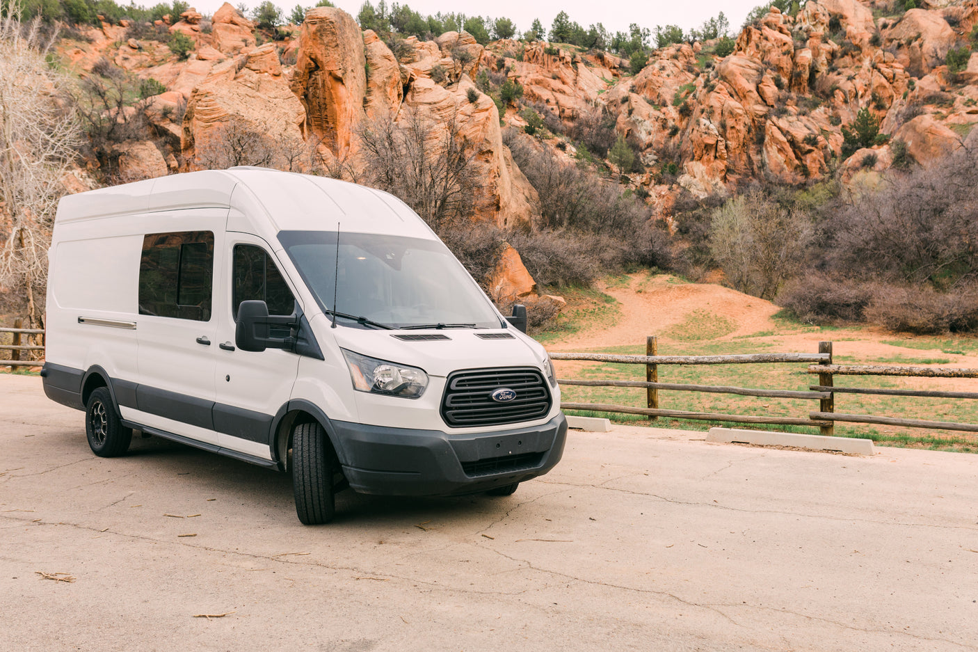 White Ford Transit RV rental parked on pavement, with striking red rock mountains and sparse trees behind.