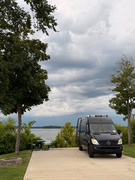 Black Mercedes Sprinter camper van at an RV rental site with a lake view under cloudy skies.