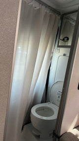 Inside an RV rental, a compact bathroom shows a white toilet with the lid open and a white shower curtain to the left.