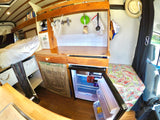 An RV rental interior showing a compact kitchen with wood counters, bed, and opened fridge with drinks.