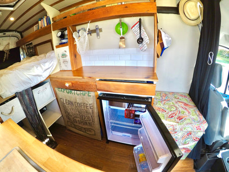 An RV rental interior showing a compact kitchen with wood counters, bed, and opened fridge with drinks.