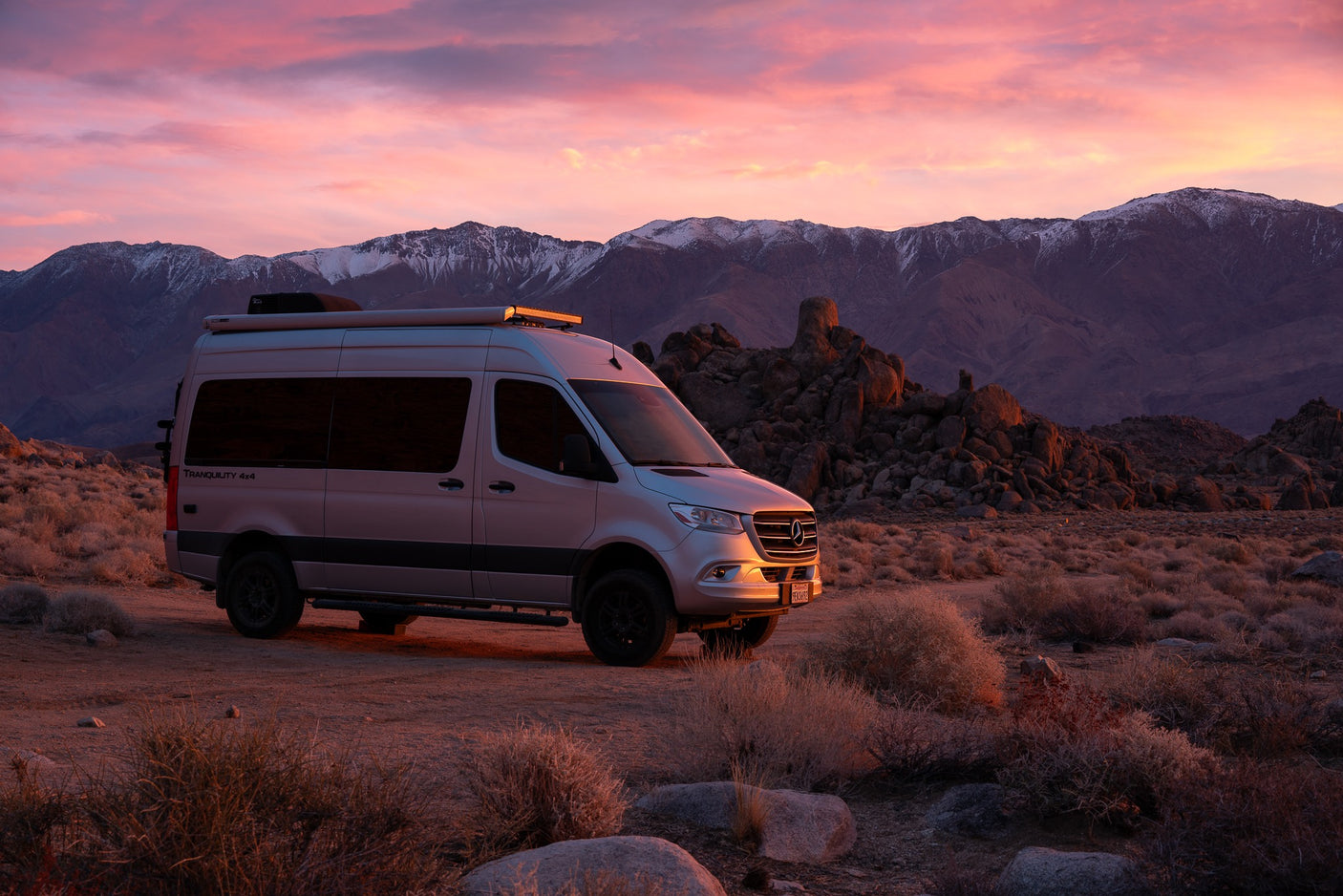 Silver Mercedes Sprinter **RV rental** at sunset. Pink sky over snow-capped mountains and a rugged desert landscape.