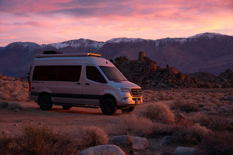 Silver Mercedes Sprinter **RV rental** at sunset. Pink sky over snow-capped mountains and a rugged desert landscape.
