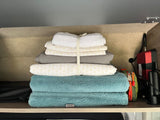 A neat stack of white and teal towels with patterned sheets, alongside travel gear, demonstrates organized RV rental storage.