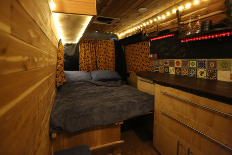 Cozy RV rental interior featuring a comfy bed, glowing wood paneling, and a colorful tiled kitchen with string lights.
