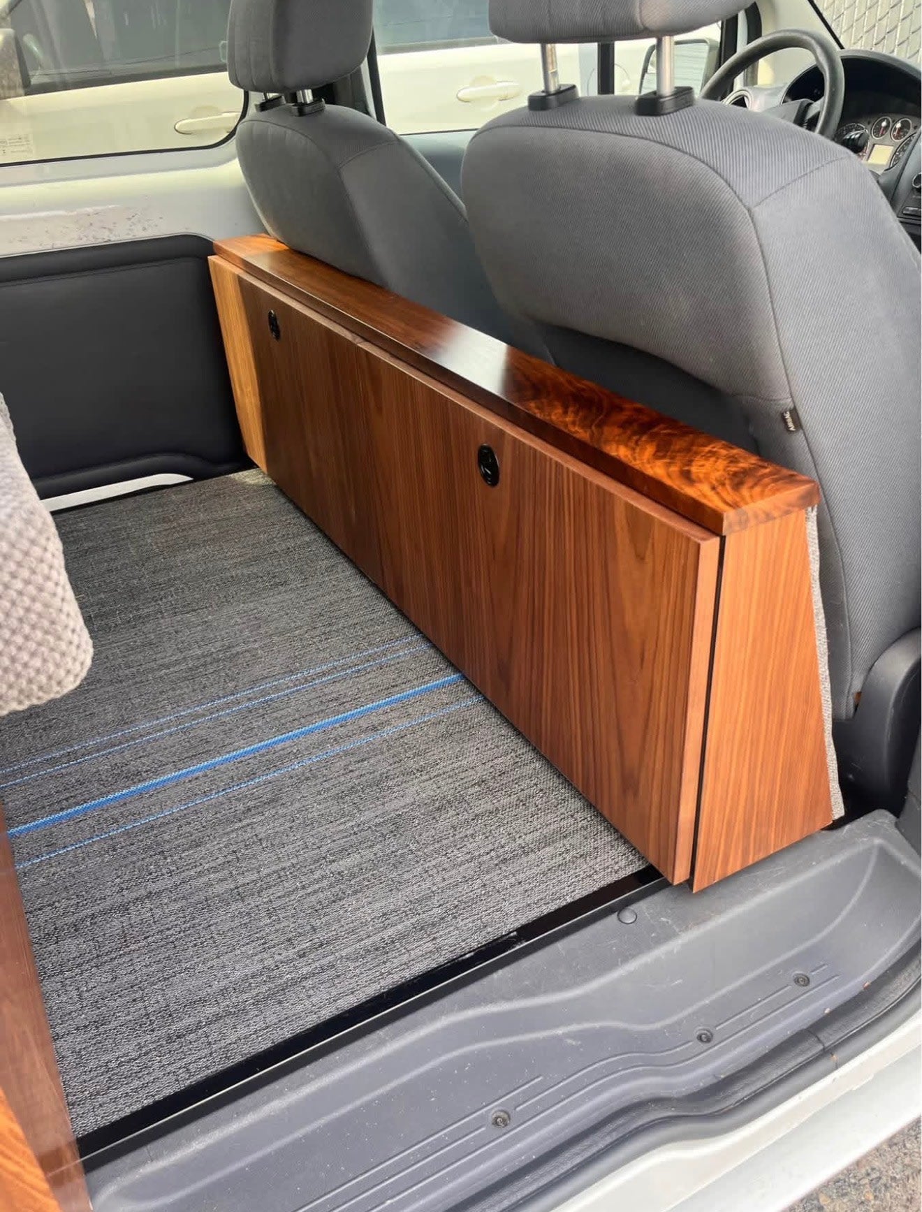  visible from the door.|RV rental interior: A comfortable grey textured bed and wooden accents inside a camper van — photo 7