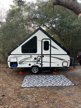 A white A-frame pop-up rv rental sits ready for camping on a pine needle forest floor with a patterned outdoor rug.