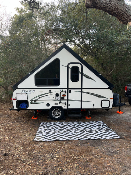 A white A-frame pop-up rv rental sits ready for camping on a pine needle forest floor with a patterned outdoor rug.