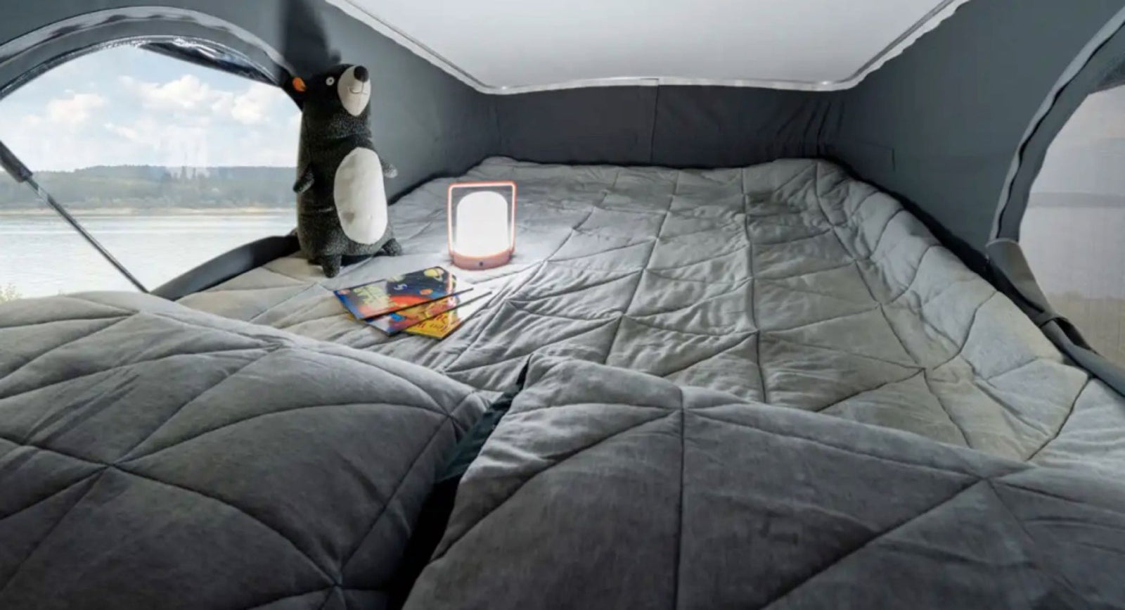 Cozy pop-top bed in an RV rental with a scenic lake view, featuring plush grey bedding, a playful bear, and a night light.