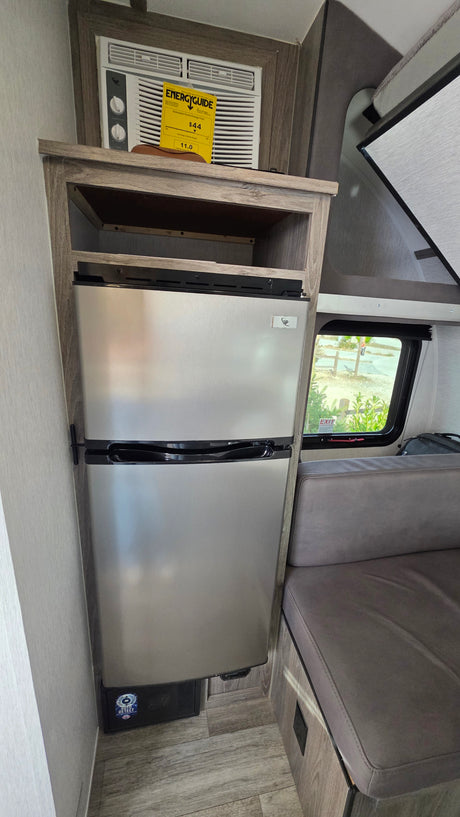 Inside an RV rental: compact stainless steel fridge, wall-mounted AC with energy label, and a window view next to seating.