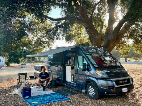 Man and dog relaxing outside their Pleasure-Way Lexor-TS RV rental (Ram ProMaster van) under a large shaded tree, enjoying outdoor camping.