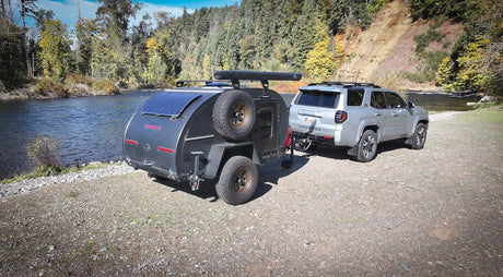 This RV rental features a silver SUV towing an off-road teardrop trailer beside a picturesque river, ideal for camping.
