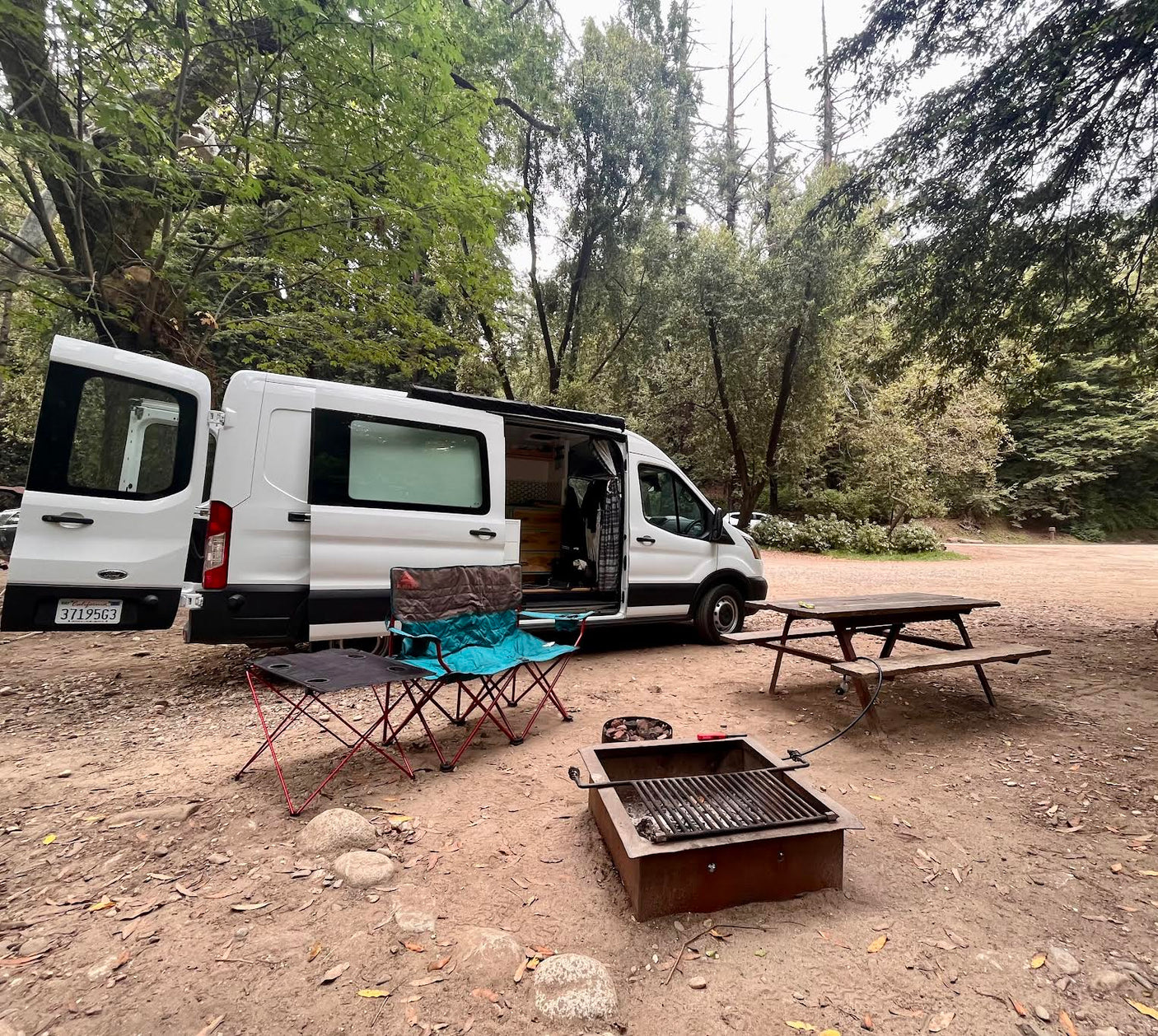 A white RV rental camper van with open doors at a forest campsite, featuring chairs, a small table, fire pit, and a picnic table.