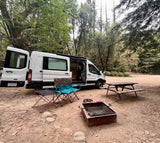 A white RV rental camper van with open doors at a forest campsite, featuring chairs, a small table, fire pit, and a picnic table.