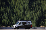 High Country Adventure RV rental van with mountain livery, parked beside a dense pine forest.