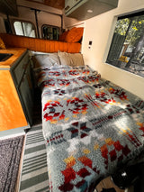 Cozy RV rental interior featuring a comfortable bed with a patterned blanket, kitchen area, and scenic window view.