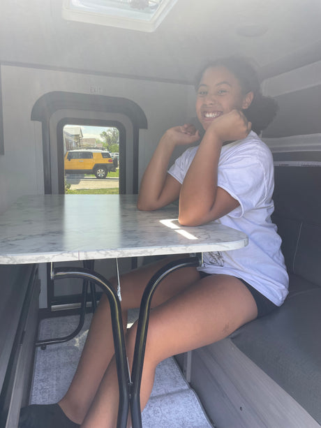 Happy girl in an **RV rental**'s cozy interior, sitting at a table looking out a window at a yellow SUV.