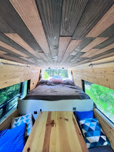Inside a unique RV rental: custom wooden ceiling & walls, cozy bed with outdoor views. Ideal for van life travel.