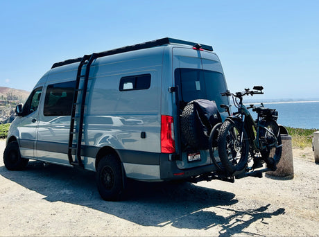Adventure-ready gray RV rental, with fat-tire e-bikes and spare, parked by the ocean.