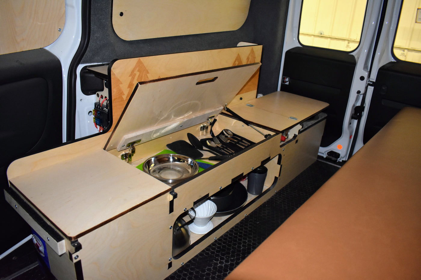 Compact RV rental interior featuring a modular wooden kitchen unit with opened lid showing cooking essentials, and a brown cushioned bed. — photo 3