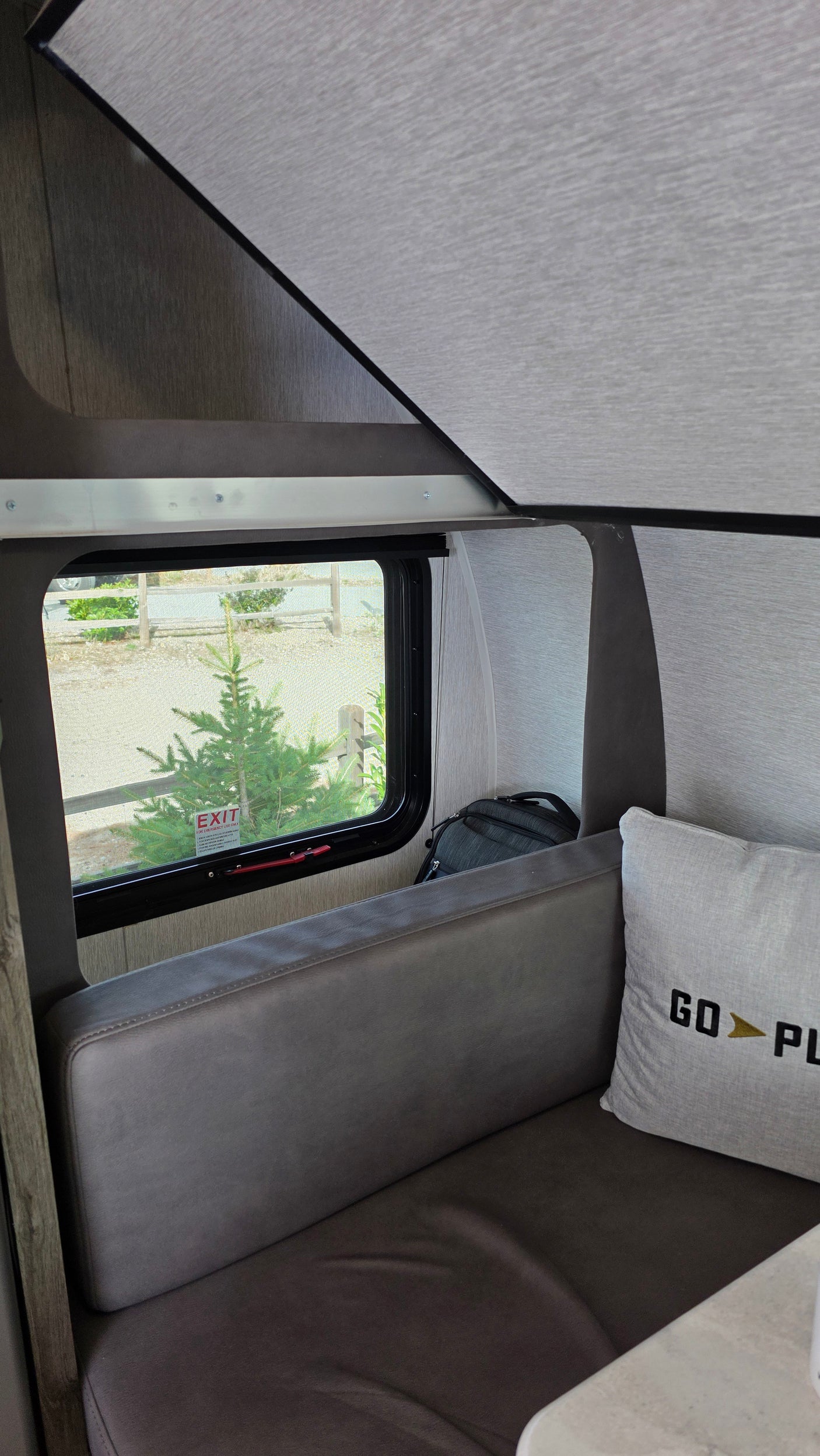 Inside an RV rental: window view of an evergreen tree and an "EXIT" sign, with a gray dinette bench and a "GO PLAY" pillow.