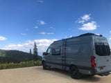 Grey Mercedes Sprinter RV rental on a mountain dirt road, surrounded by wildflowers & pines under blue sky.