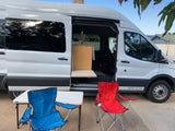 White RV rental van with open side door, showing interior kitchenette; outdoor seating setup with chairs and table.