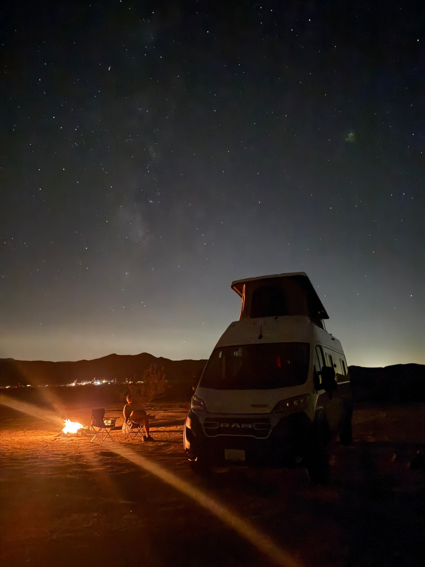  parked in a vast desert landscape at sunset.|A white RV rental camper van with a pop-top roof is set up for desert camping at sunset. Chairs await.|White Winnebago Solis RV rental van parked in a quiet desert setting with mountains at sunset.|Serene desert camping with a white RV rental. Pop-top extended — photo 16