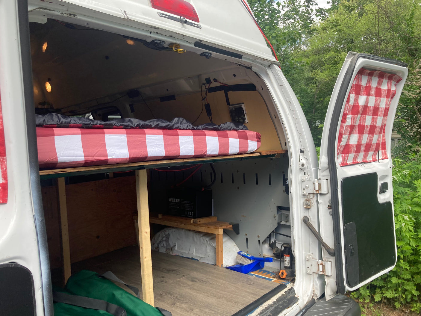 Interior of a converted RV rental van, featuring a cozy bed with red/white checkered bedding, under-bed storage, and power setup.