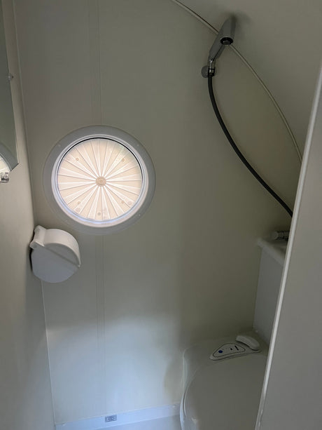 RV rental bathroom features: a circular skylight with a pleated blind, a compact toilet, and a wall-mounted shower head.
