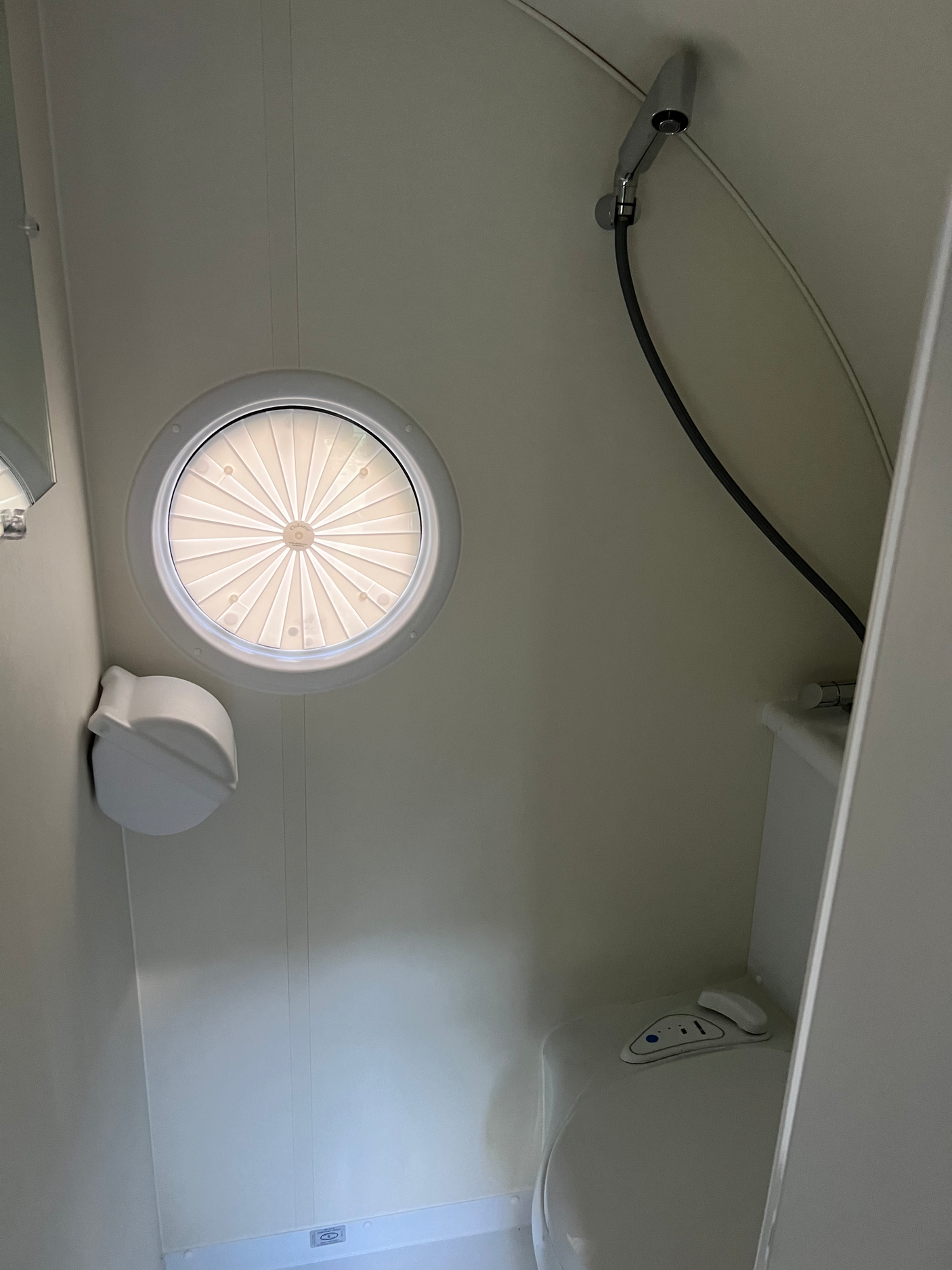 RV rental bathroom features: a circular skylight with a pleated blind, a compact toilet, and a wall-mounted shower head.
