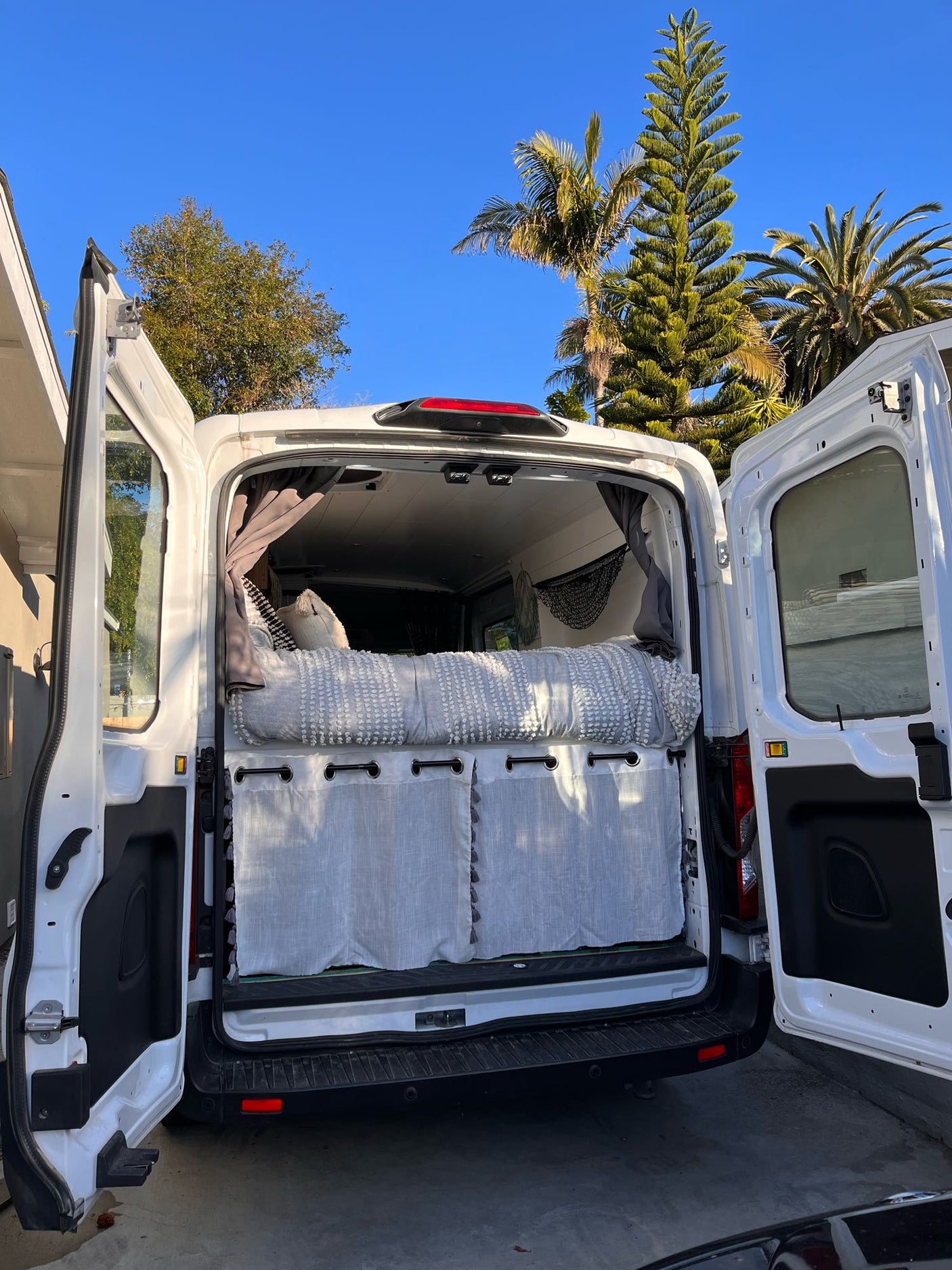 White camper van (rv rental) with open rear doors, showing a cozy bed, gray bedding, & privacy curtains inside. Sunny day, palm trees.