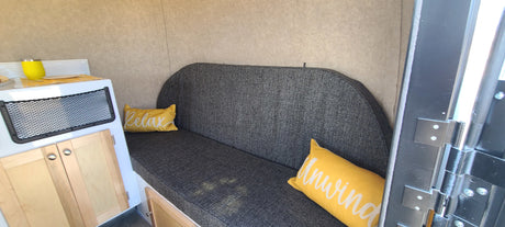 Compact RV rental interior: gray upholstered bench with 'Relax' & 'Unwind' pillows, light wood cabinets, and built-in storage net.