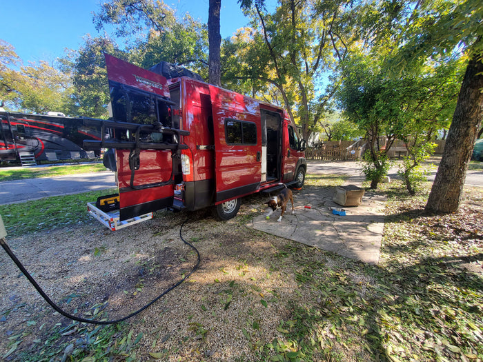 Red RV rental camper van parked at a sun-dappled campsite, with a playful dog on the ground.