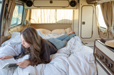 Woman relaxes in an RV rental, reading a book on a striped bed inside a cozy camper van interior.