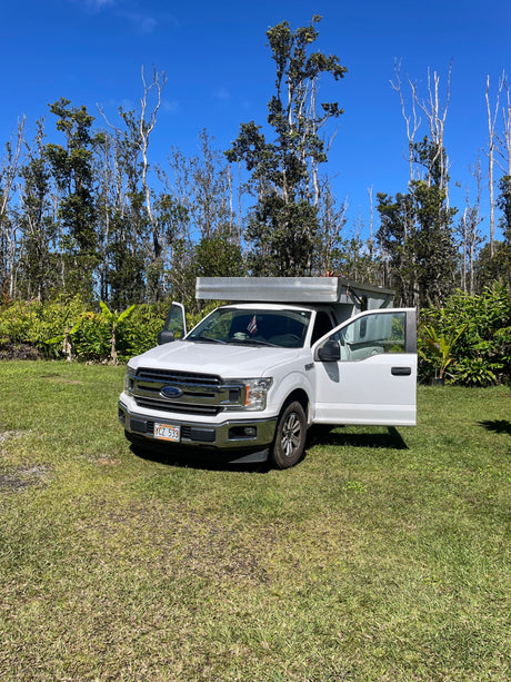 White pickup truck with camper, ready as an RV rental, parked on grass with doors open under a blue sky, surrounded by trees.