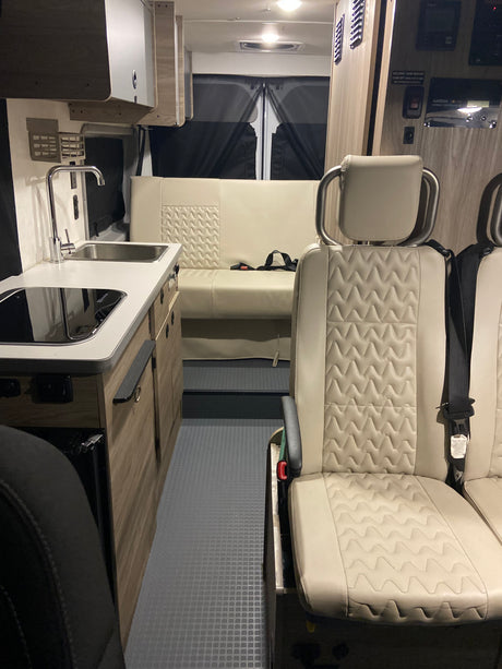 Experience an RV rental! View of a modern camper van interior with quilted beige seats, a compact kitchen, and rear bench.