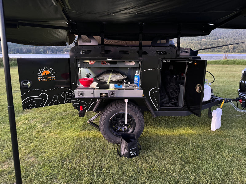 This off-grid trailer for rv rental features an open side revealing a well-equipped outdoor kitchen, parked on grass beside a tranquil lake. — photo 6