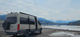 An RV rental, a customized Mercedes Sprinter van, parked on a riverbank with mountains, a bridge, and other RVs in the background.