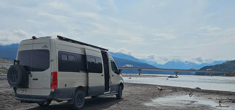 An RV rental, a customized Mercedes Sprinter van, parked on a riverbank with mountains, a bridge, and other RVs in the background.
