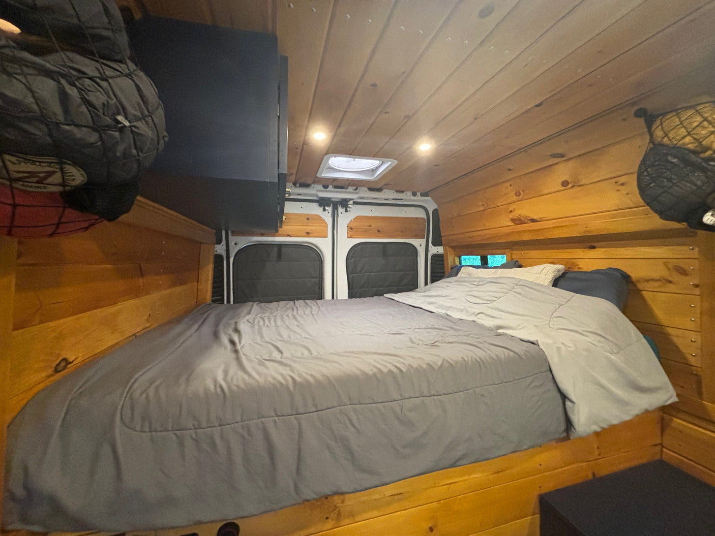 Experience this cozy rv rental! Van interior features a comfortable bed, wooden walls, ceiling lights, and overhead storage.