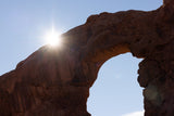 A bright sunburst peeks over a massive sandstone arch against a blue sky, an ideal stop for an RV rental trip.