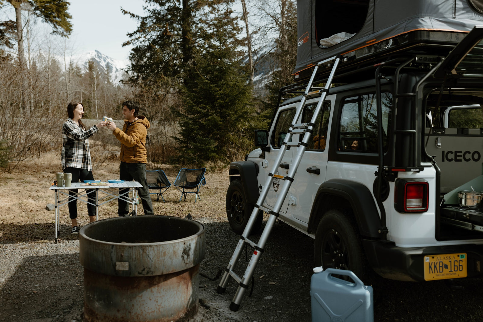 Experience outdoor adventure with an RV rental! A couple enjoys a toast at their Alaska campsite, complete with a Jeep & rooftop tent.