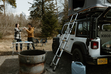 Experience outdoor adventure with an RV rental! A couple enjoys a toast at their Alaska campsite, complete with a Jeep & rooftop tent.