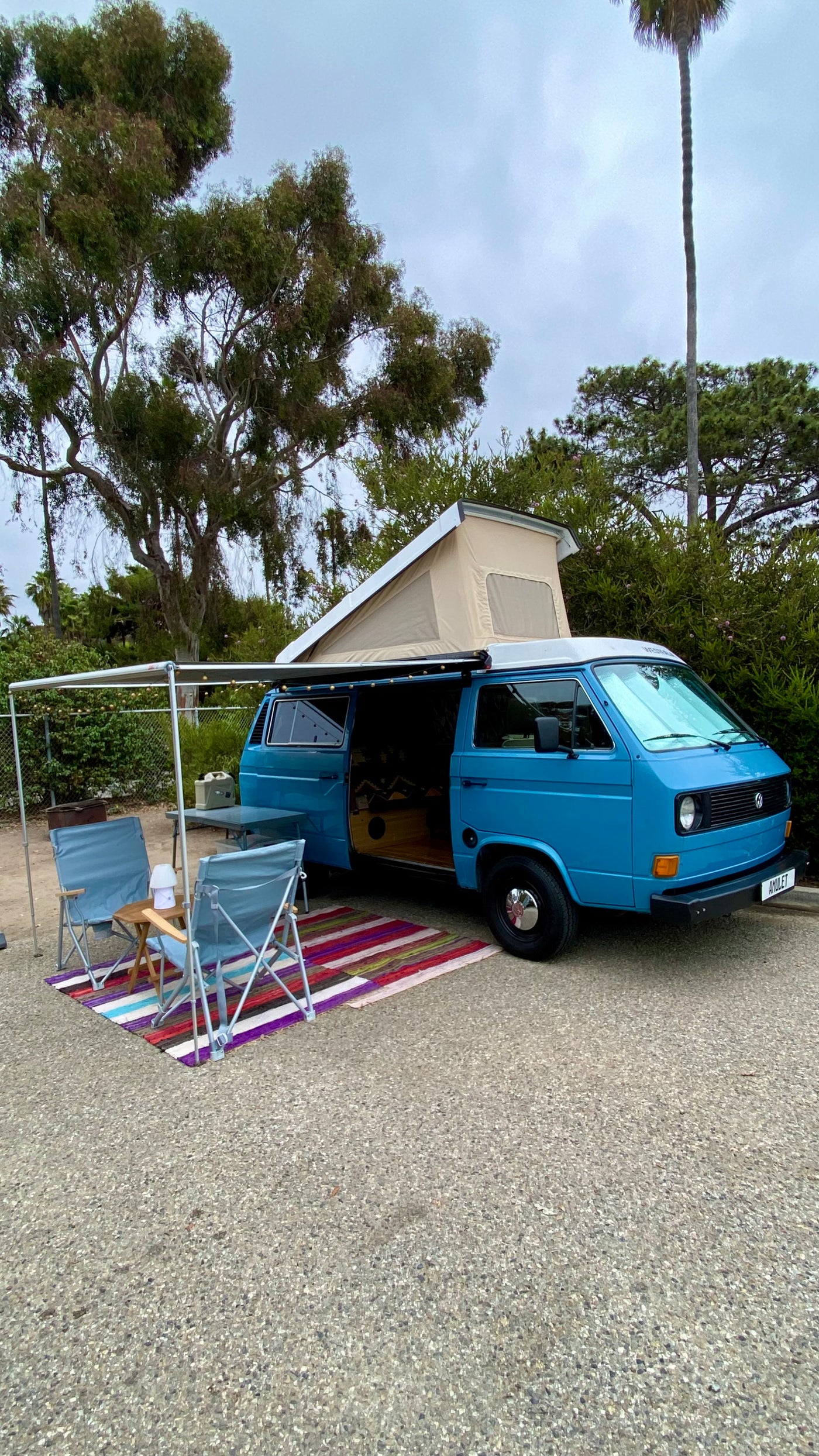 An inviting blue camper van, ideal for an rv rental, with its pop-top extended, awning, chairs, and colorful rug set up.
