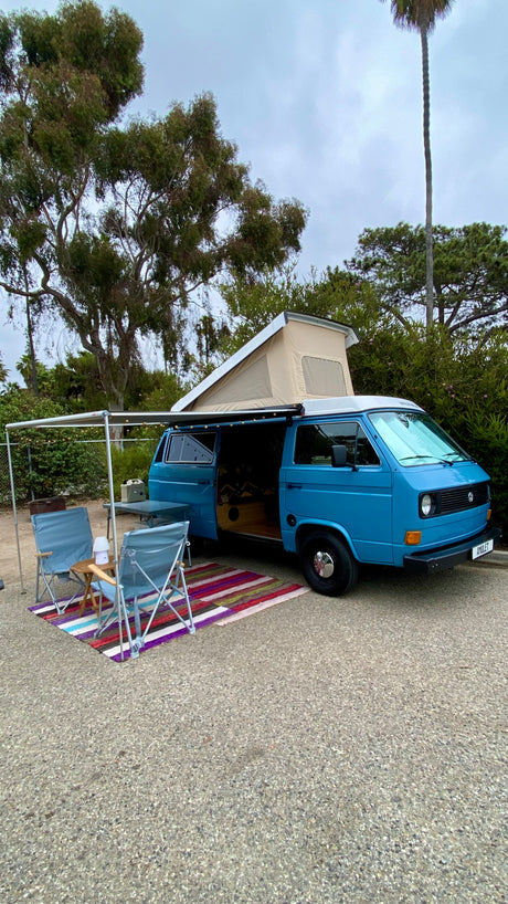 An inviting blue camper van, ideal for an rv rental, with its pop-top extended, awning, chairs, and colorful rug set up.