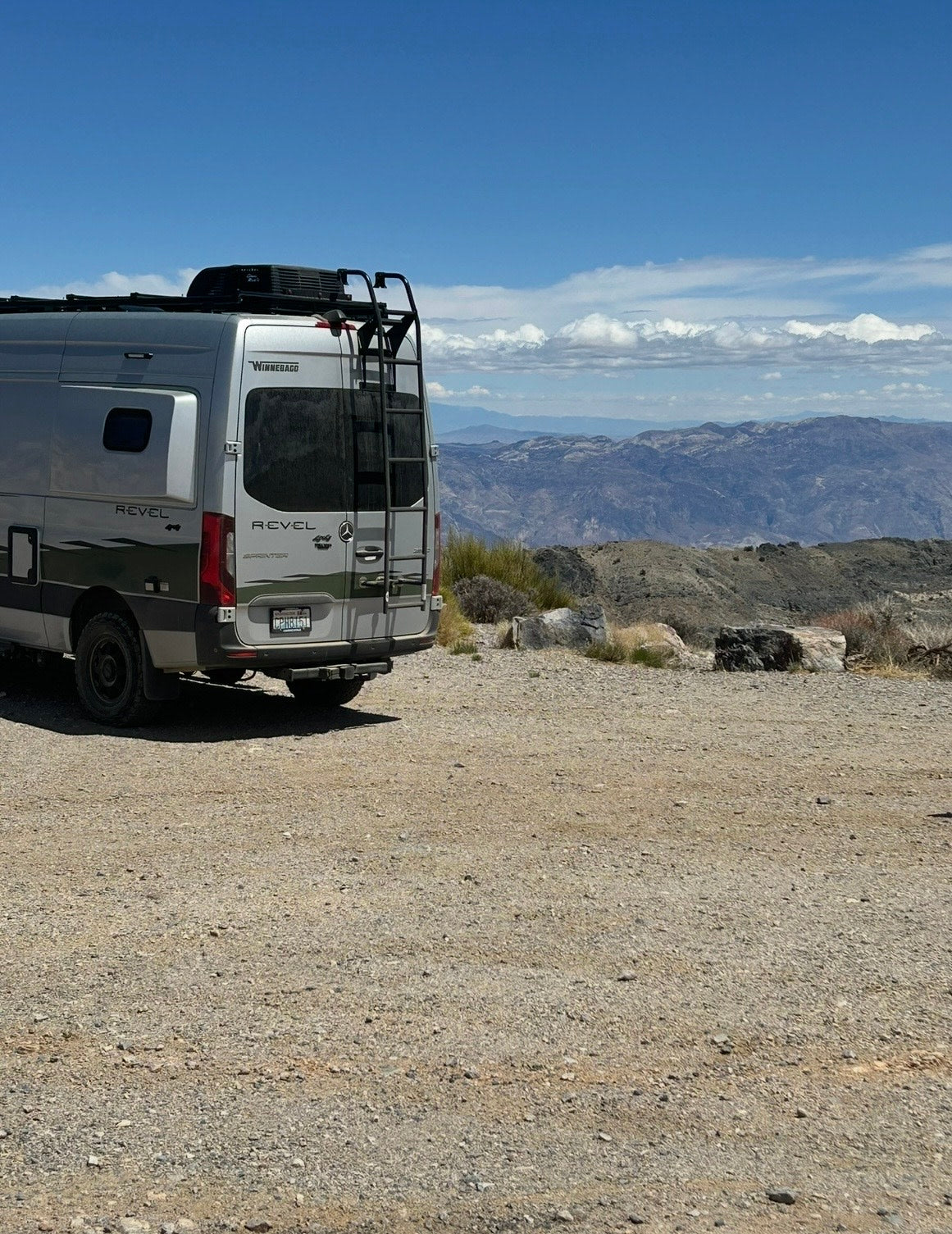 Winnebago Revel RV rental parked on gravel with ladder & roof rack, overlooking a vast mountain landscape under a blue sky.
